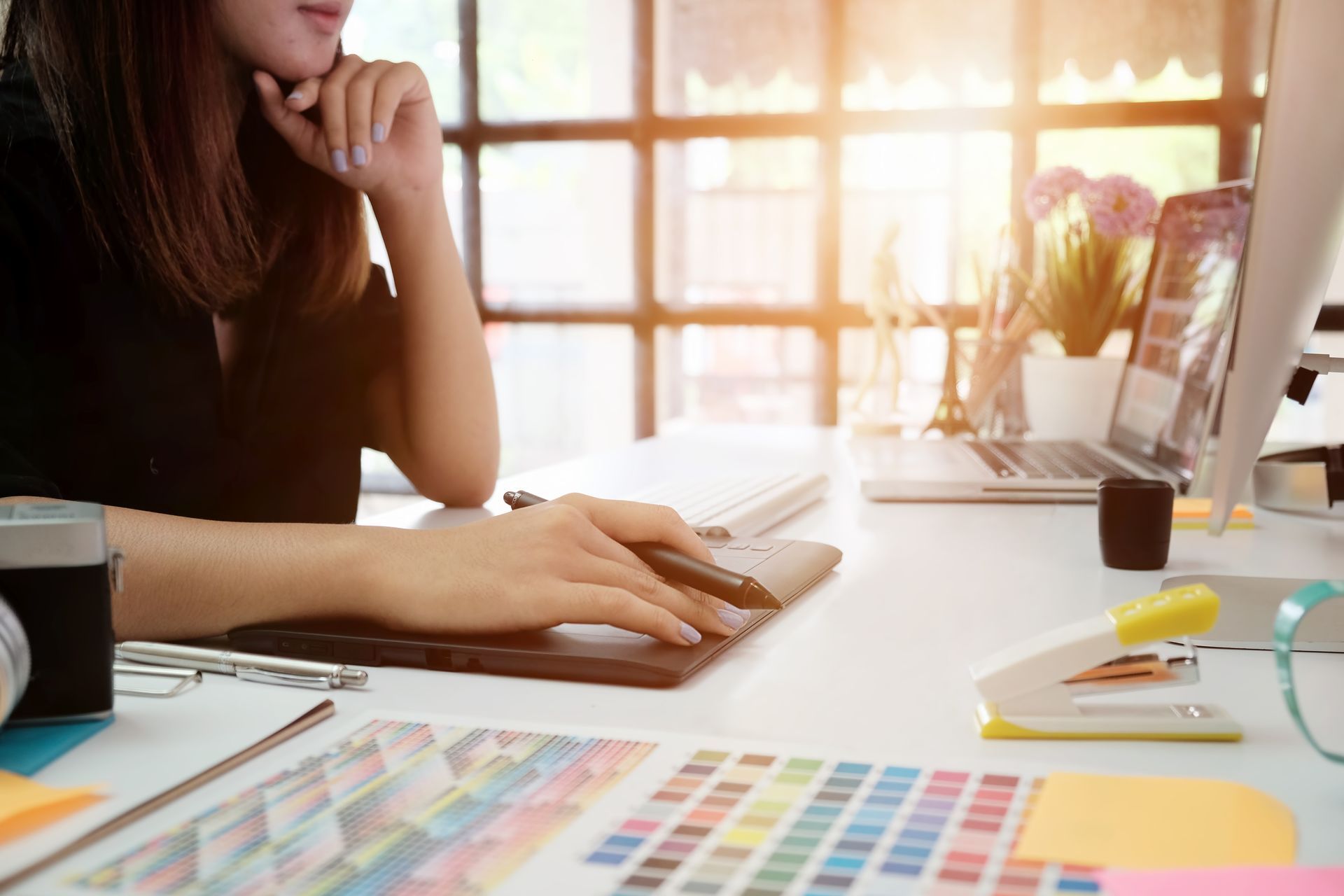 Une personne travaille à un bureau avec des échantillons de couleurs, un ordinateur portable et la lumière du soleil entrant par une fenêtre.