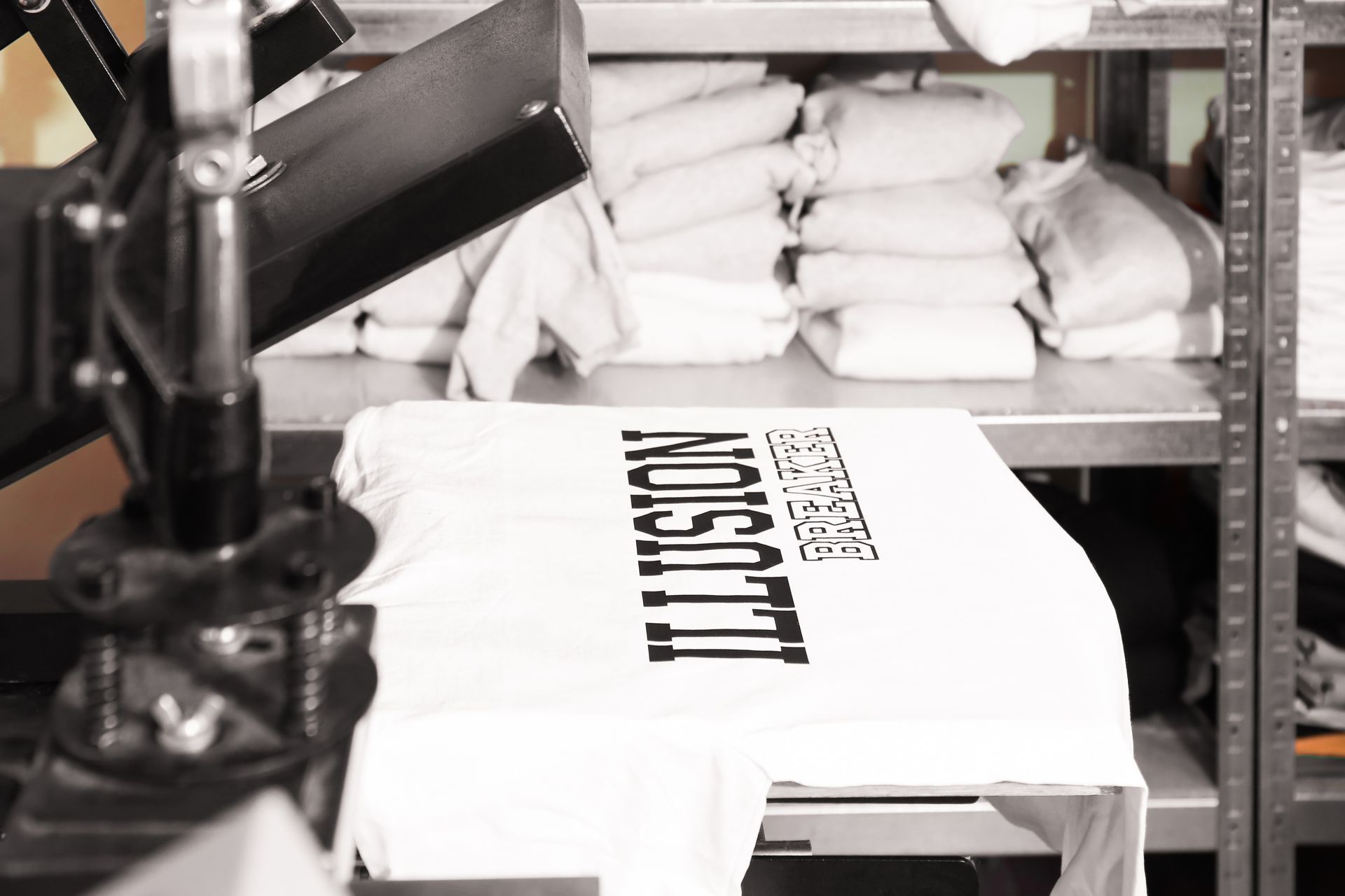 Des t-shirts sont imprimés sur une presse dans un atelier de confection, avec des t-shirts pliés sur des étagères derrière.