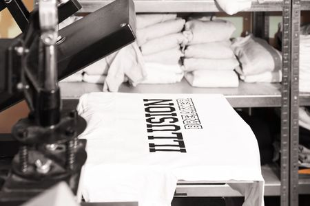 Un t-shirt blanc imprimé est posé sur une table de production, avec des t-shirts pliés sur des étagères derrière.