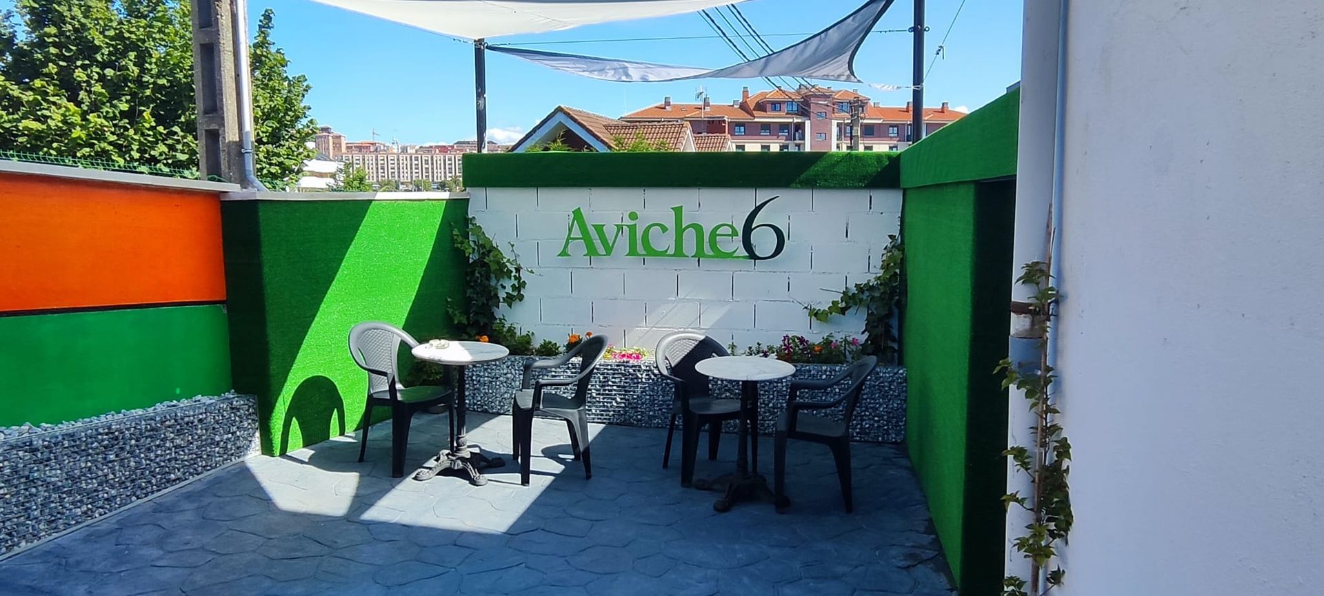 Zona de estar al aire libre con paredes verdes, cartel blanco "Aviche6", mesas y cielo azul.