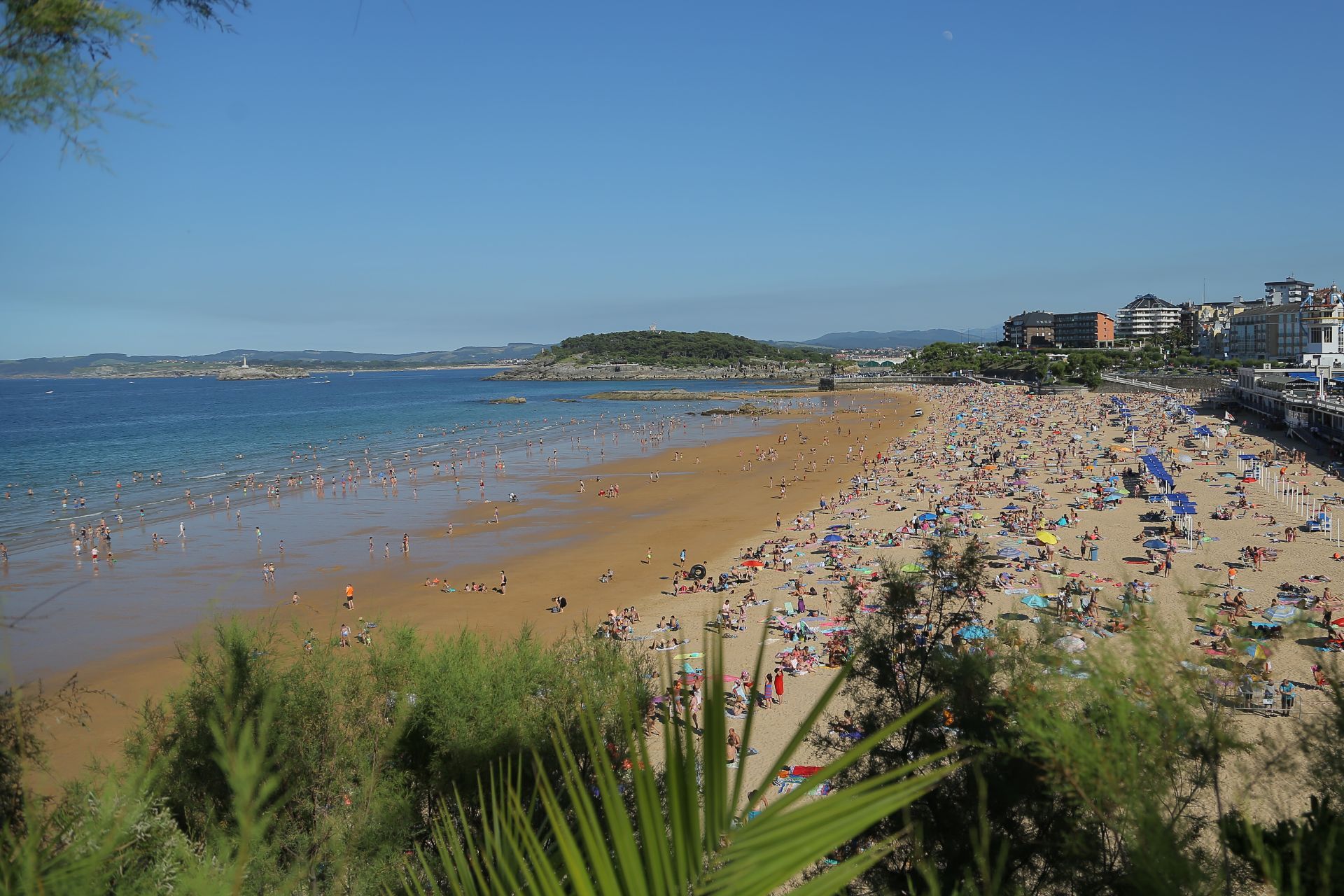 Playa llena de gente bajo un cielo azul despejado. A lo lejos se ven edificios y una pequeña isla.