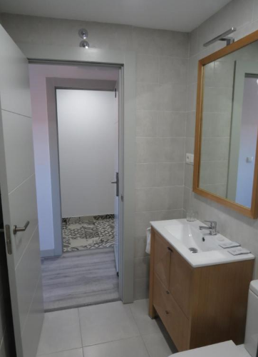Baño con puerta abierta, tocador de madera clara, espejo cuadrado, azulejos grises y piso blanco.