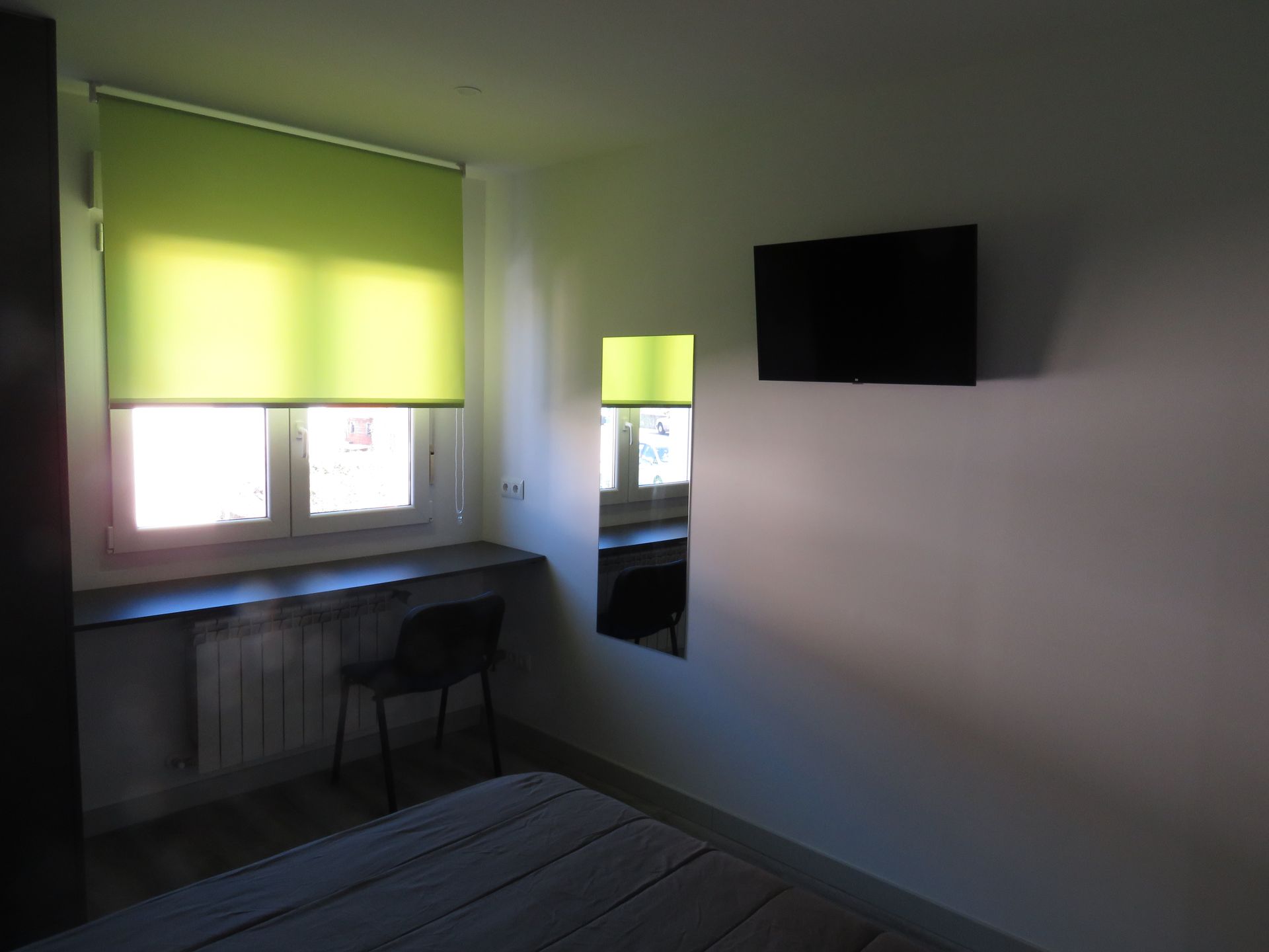Dormitorio con ventana, escritorio, silla, espejo y televisor en la pared. Persiana enrollable verde.