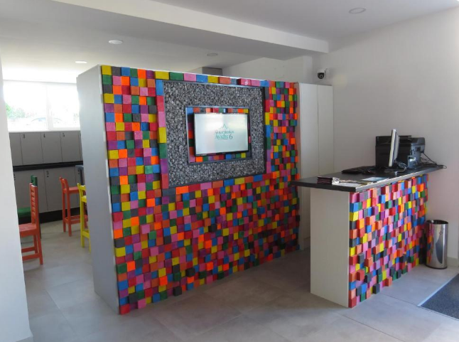 Mostrador de recepción con pared de bloques de colores, televisor y computadora. Zona de espera visible.