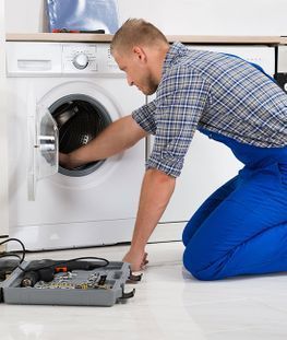 Una persona con un mono azul repara una lavadora, arrodillada y con una caja de herramientas cerca.