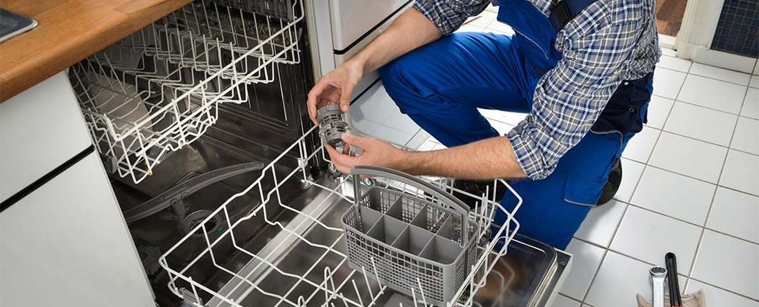 Persona con ropa de trabajo azul reparando un lavavajillas, sosteniendo vasos cerca de la cesta de cubiertos.