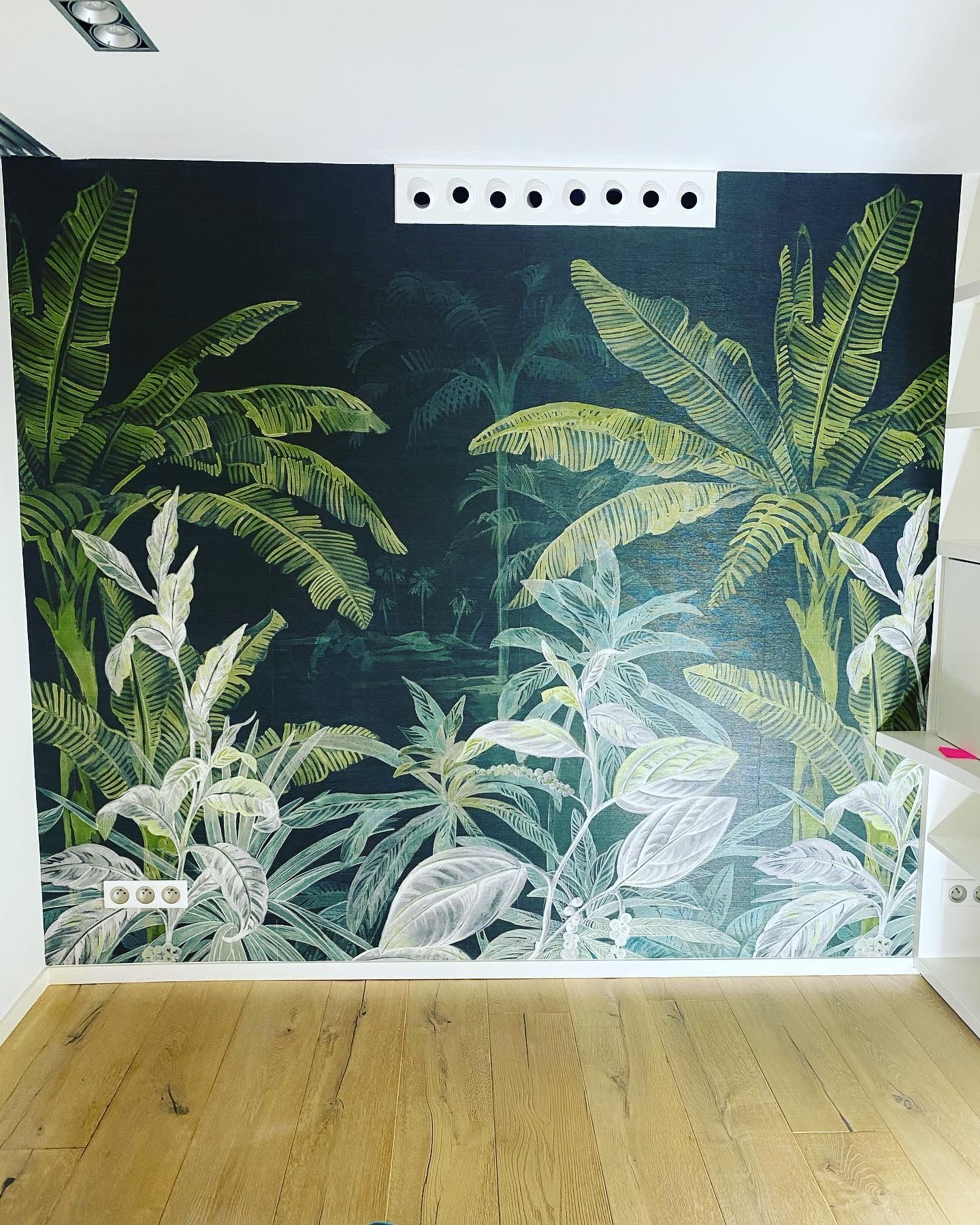 Peinture murale sur le thème de la jungle sur fond sombre ; feuillage vert et blanc. Mur blanc avec étagères intégrées ; parquet.