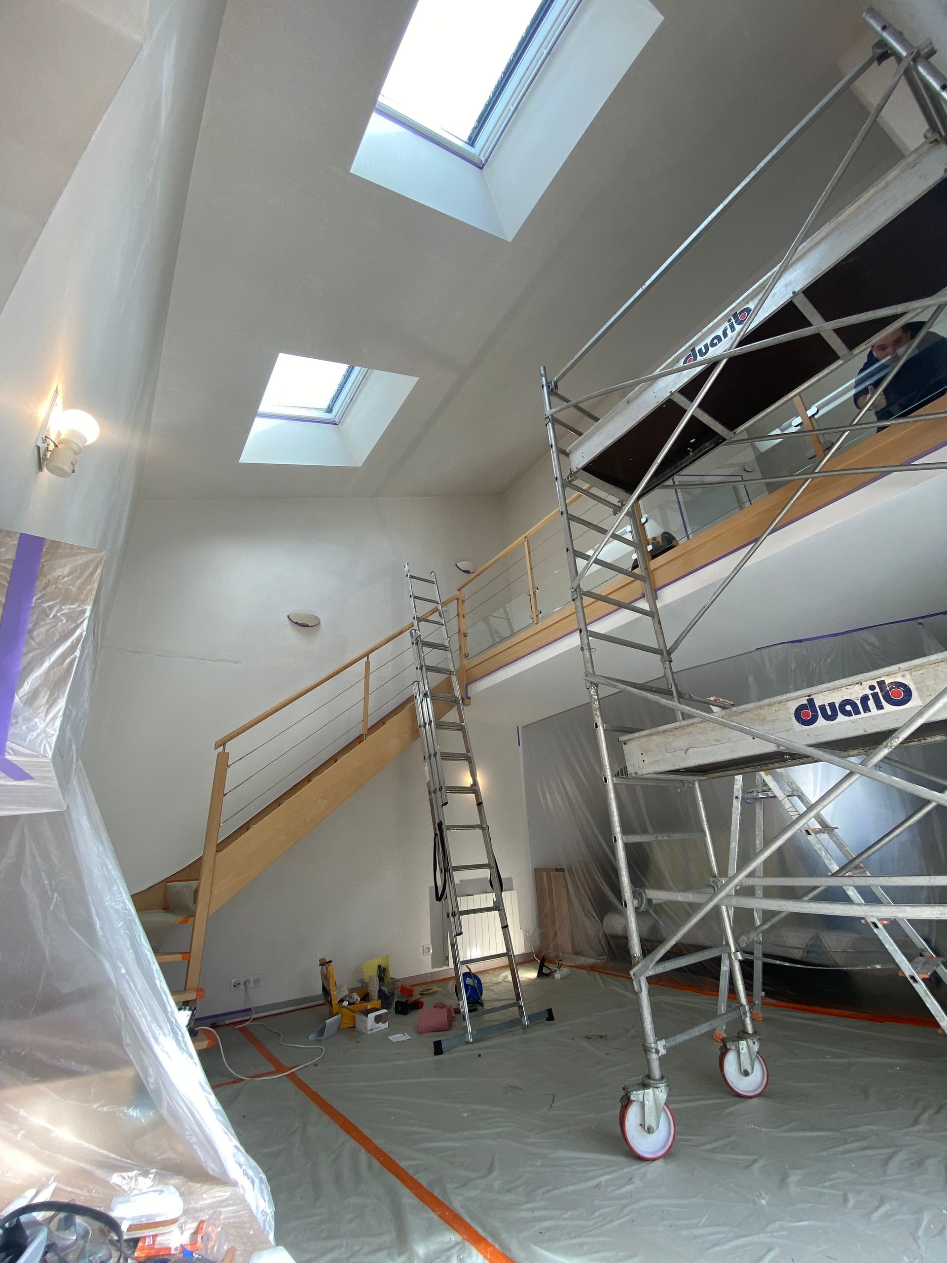 Vue intérieure d'un chantier avec échafaudages, escaliers et puits de lumière. La pièce est recouverte de plastique.