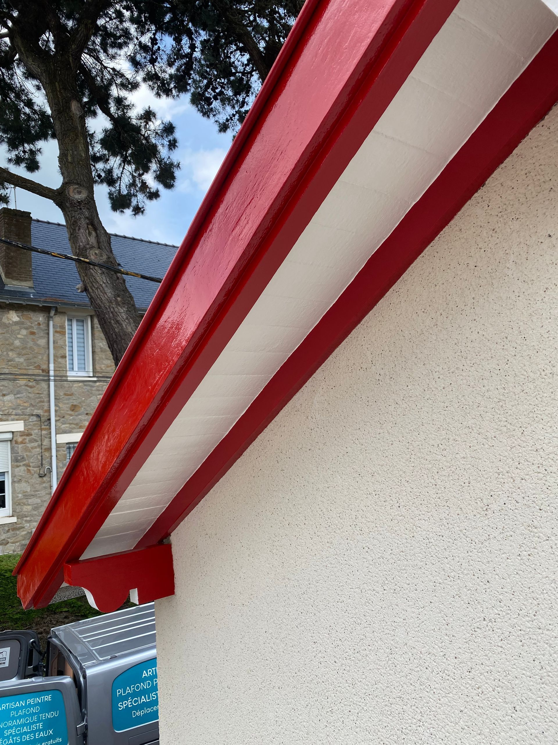 Avant-toit peint en rouge avec sous-face blanche sur un bâtiment en stuc. Un arbre et un bâtiment gris apparaissent en arrière-plan.