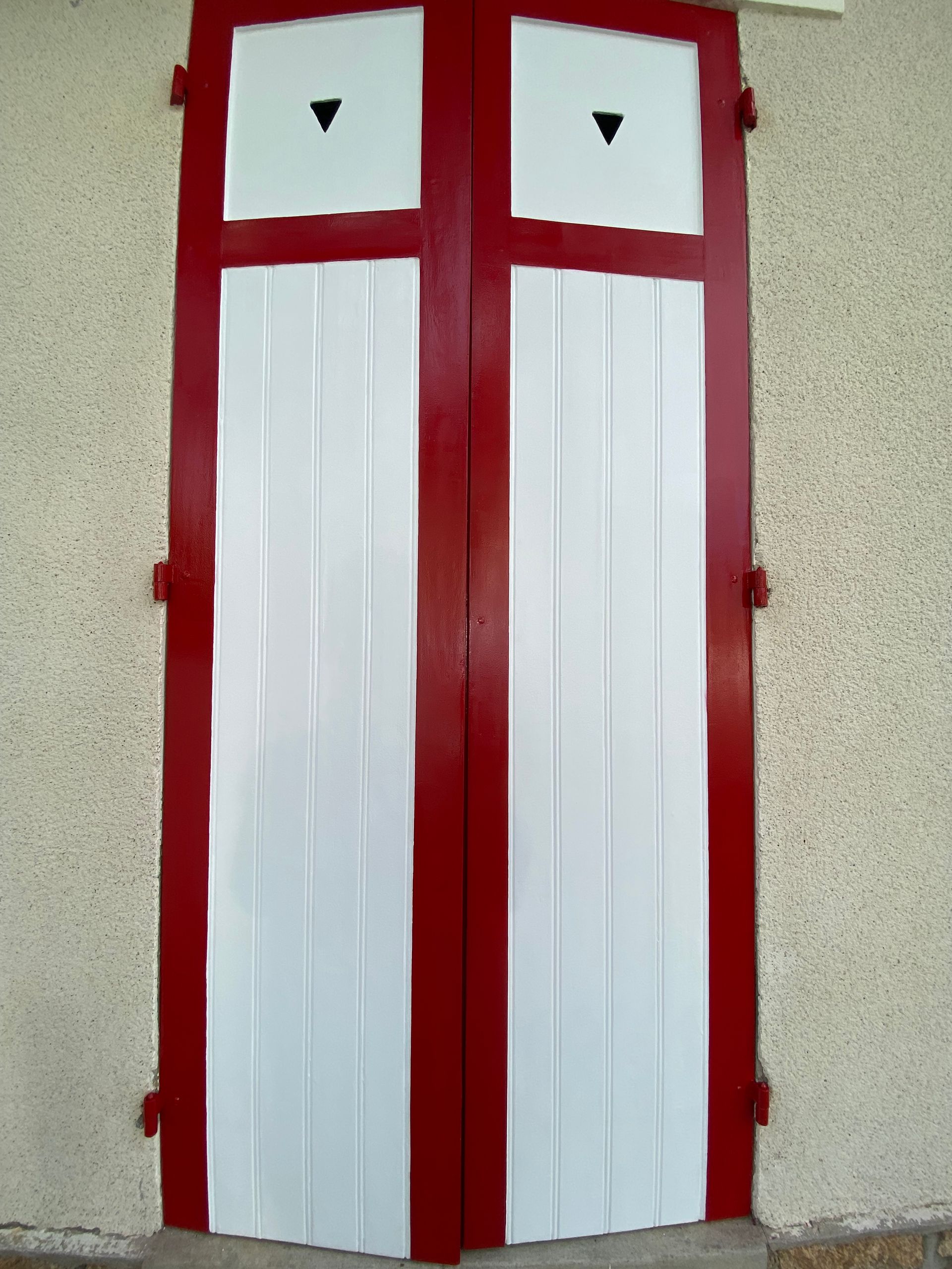 Volets blancs à cadre rouge sur un mur en stuc. Chaque volet est doté d'un carré blanc surmonté d'un triangle noir.