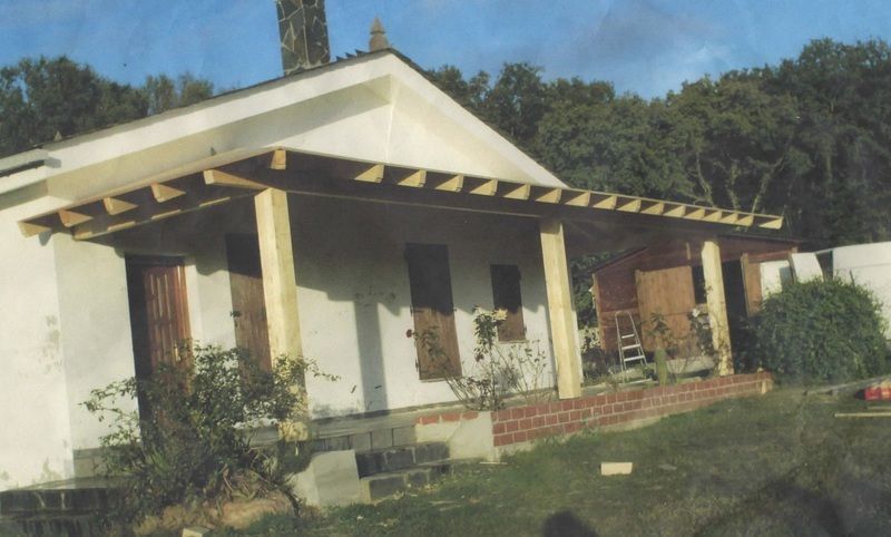 Casa blanca con un porche en construcción, sostenido por vigas de madera. La vegetación rodea la estructura.