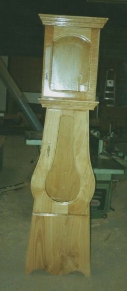 Un reloj de pie alto de madera en un taller. El reloj es de color marrón claro con la parte inferior redondeada.