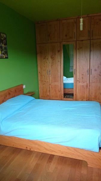 Un dormitorio con cama de madera y armario. Pared verde, ropa de cama azul y suelo de madera.