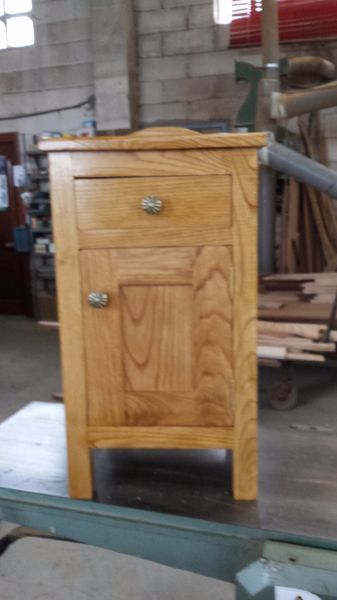 Mesita de noche de madera con cajón y puerta, teñida en marrón claro, situada en un taller.