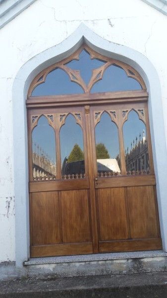Puertas dobles de madera con parte superior arqueada y paneles de vidrio ornamentados; entorno al aire libre.