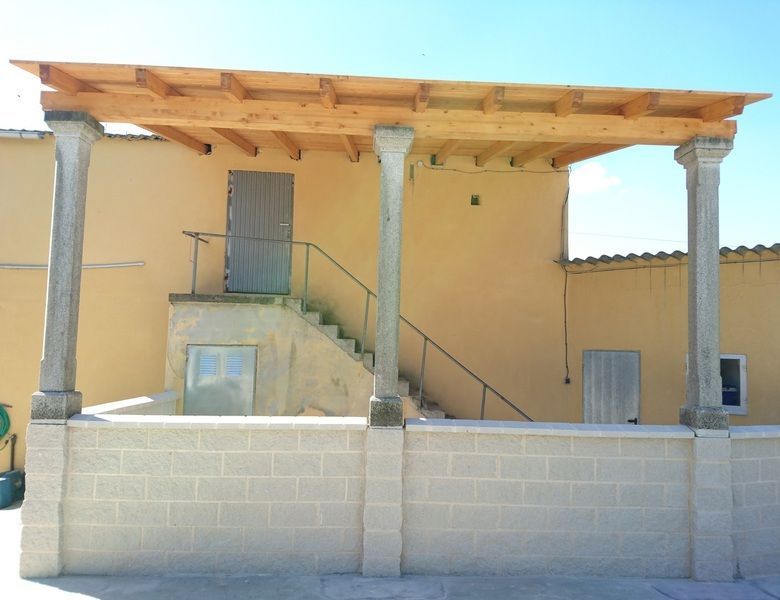 Una pérgola con techo de madera sostenida por columnas de piedra frente a un edificio amarillo.