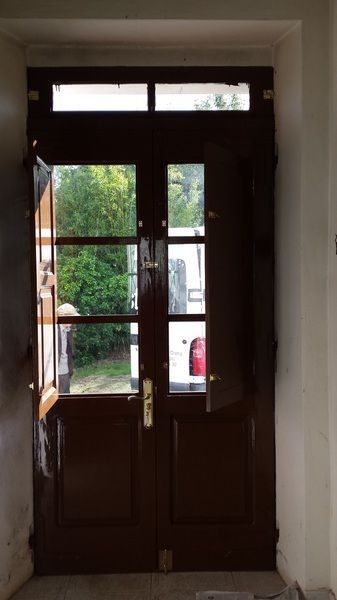 Puertas dobles de madera de color marrón oscuro, una abierta, con paneles de vidrio en la parte superior, que revelan una vista de árboles y un edificio.