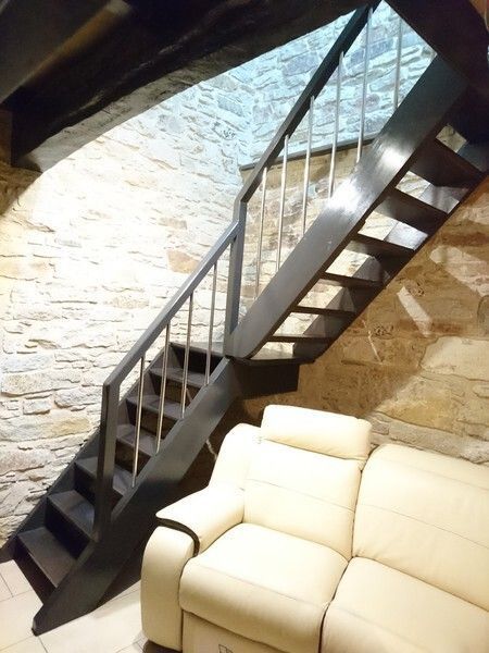 Una escalera con escalones de color gris oscuro y barandillas de metal conduce desde un sofá de cuero blanco, apoyado contra una pared de piedra.