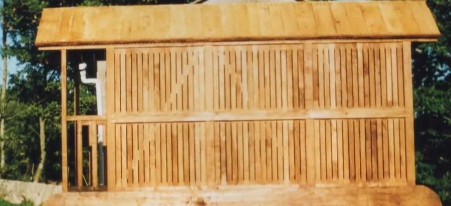 Estructura exterior de madera con paredes de listones verticales, cubierta sencilla y zona de porche, rodeada de vegetación.