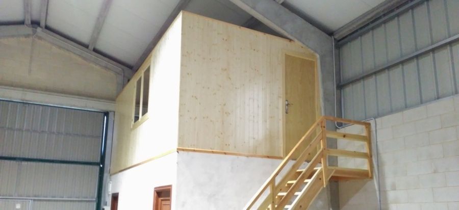 Vista interior de un entrepiso de madera de nueva construcción en una nave metálica. Una escalera conduce a una puerta y una ventana.