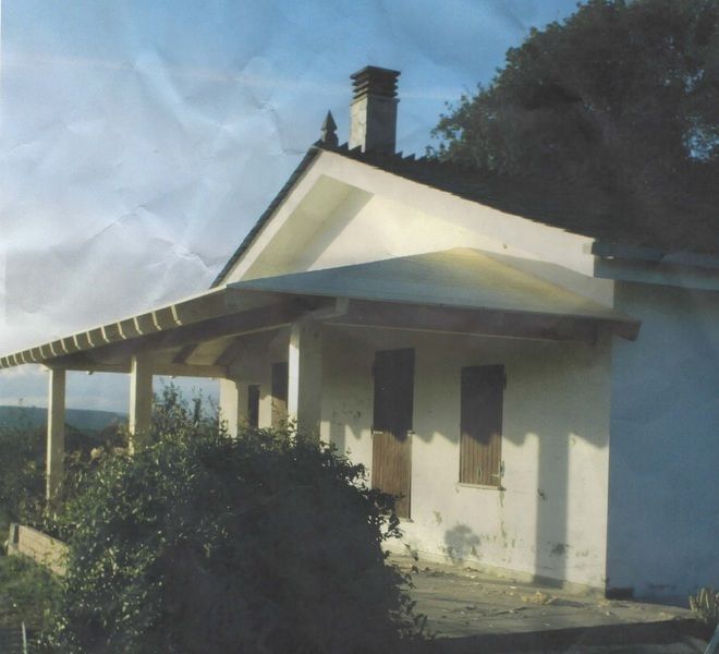 Edificio blanco de una planta con porche, persianas y chimenea. Cielo nublado. Arbustos frente al edificio.