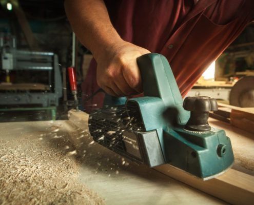Persona que utiliza una cepilladora eléctrica manual de color verde azulado para alisar una tabla de madera en un taller.