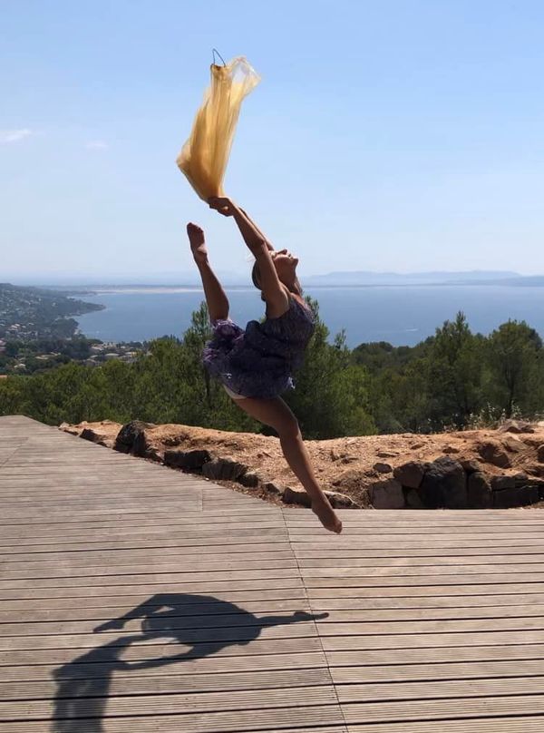 Danseuse sautant avec un tissu jaune, fendu en plein air sur une terrasse en bois avec vue sur l'océan.