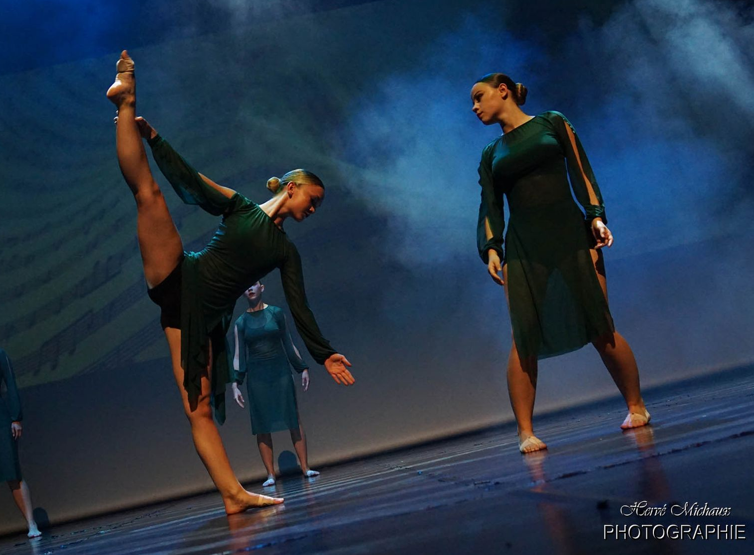Des danseurs en robes vertes se produisent sur scène, une jambe tendue vers le haut, les autres en arrière-plan.