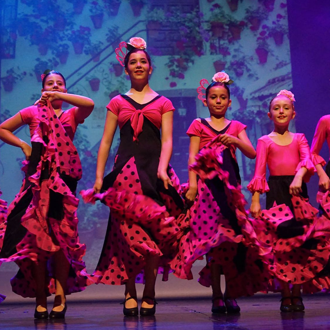 Danseuses en robes de flamenco à pois roses sur scène, avec un fond de fleurs, tenant leurs jupes.