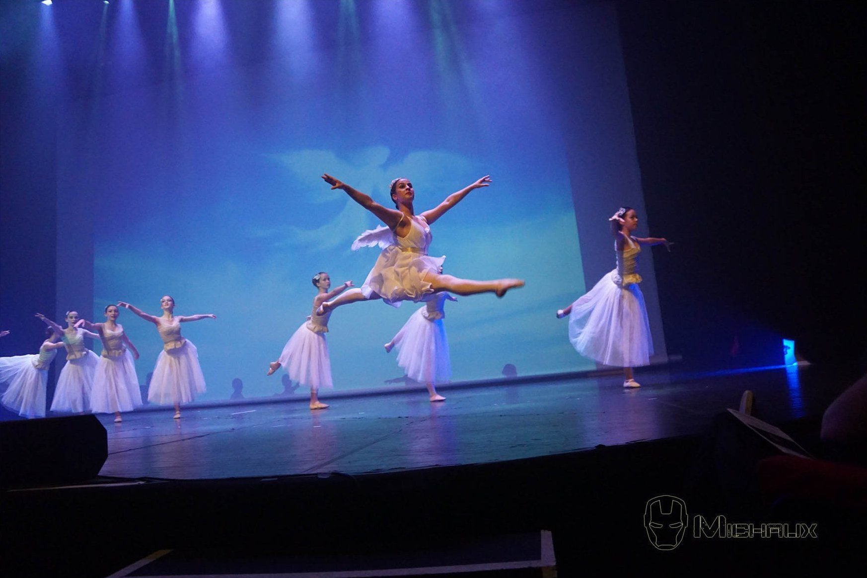 Des danseurs de ballet se produisent sur scène ; l'un en plein vol, d'autres en tutus blancs, éclairage de scène bleu.