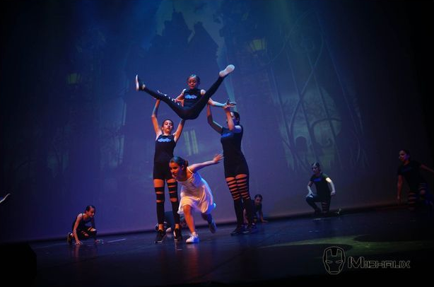 Des danseurs exécutent une routine scénique. L'un d'eux est soulevé, les autres soutenus en bas. Costumes sombres, fond bleu.