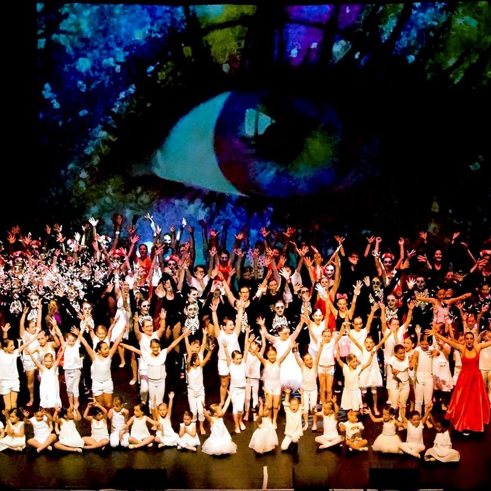 Grand groupe sur scène, bras levés, sous une projection oculaire géante. Beaucoup sont vêtus de blanc. Scène sombre.