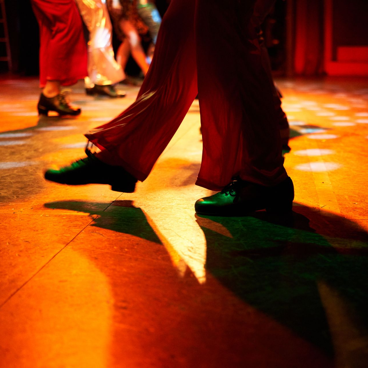 Les jambes des danseurs en tenues rouges et vertes se déplacent sur une piste de danse en bois orange brillant.