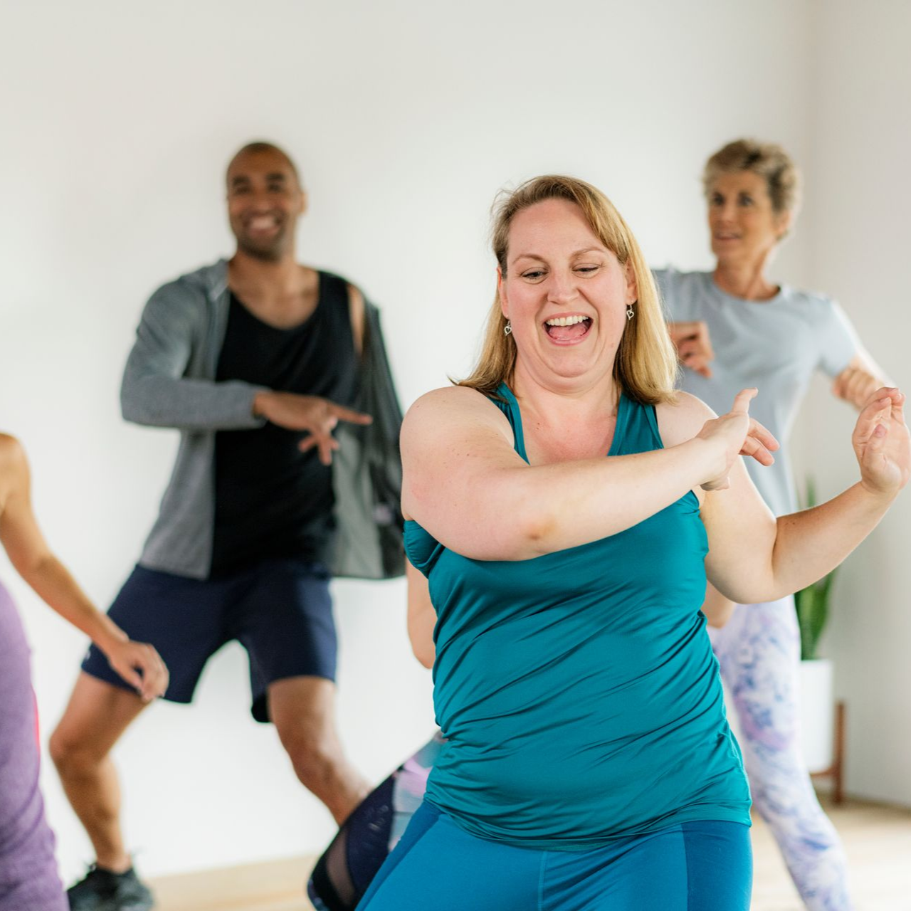 Des gens dansent dans un studio lumineux. Une femme en chemise bleu canard rit en bougeant les bras. D'autres dansent aussi.