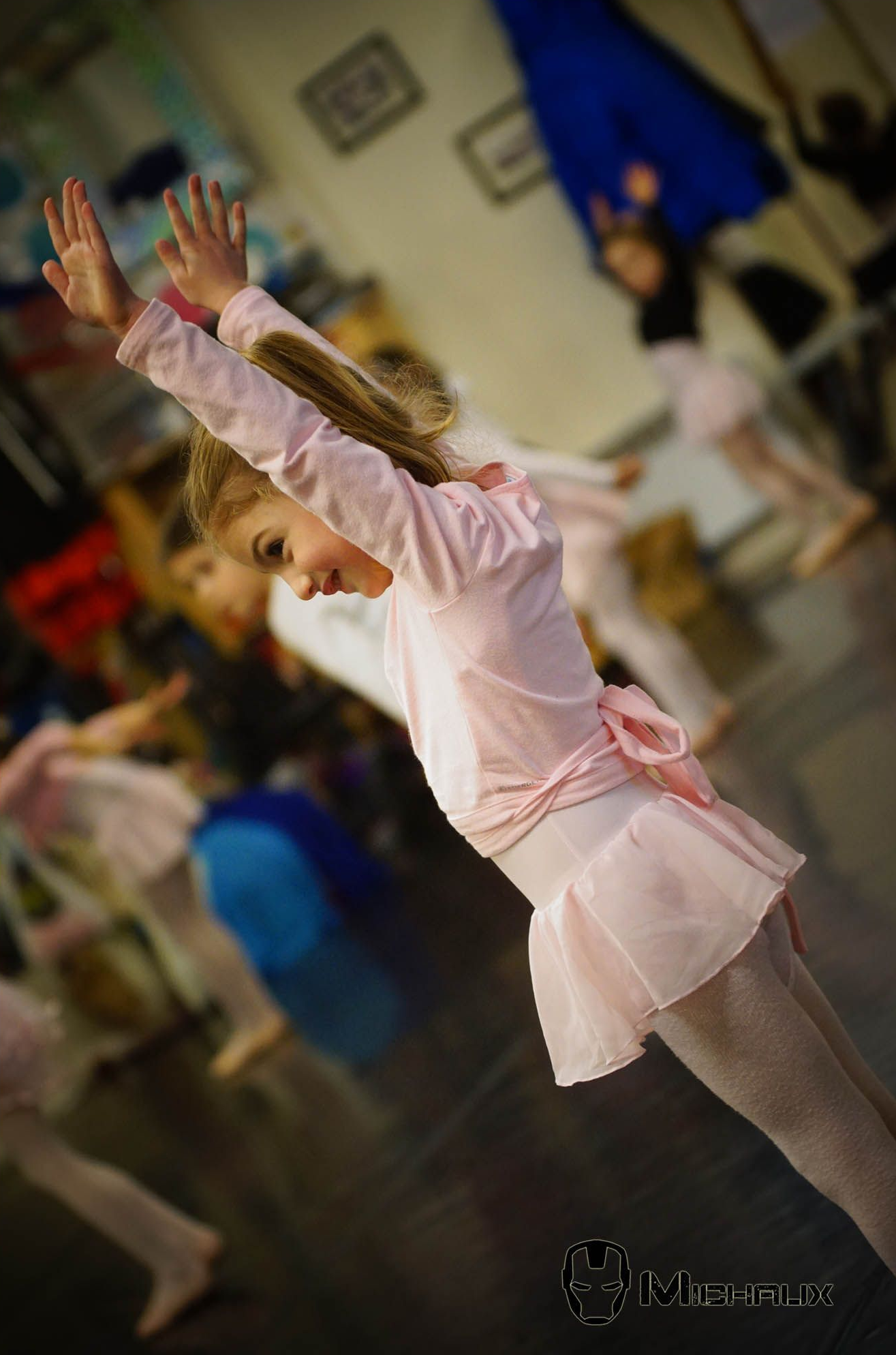 Fille en tenue de ballet rose, les bras levés, dans un studio de danse avec d'autres.