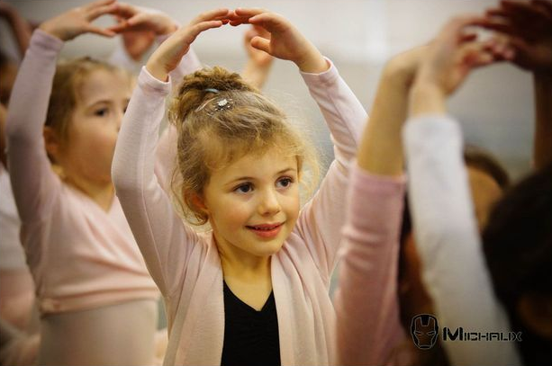 Jeune fille en cours de danse classique, les bras au-dessus de la tête, souriante. D'autres personnes à l'arrière-plan.