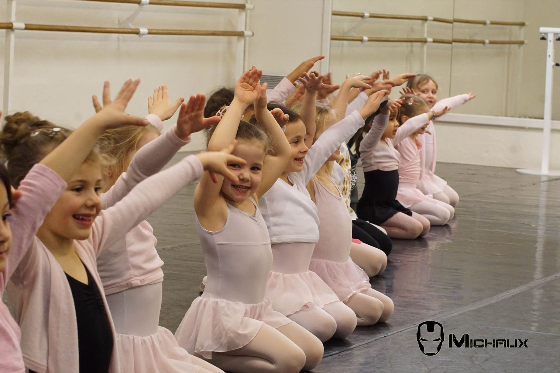 Jeunes danseurs en tenue rose agenouillés, les bras levés dans un studio de danse.