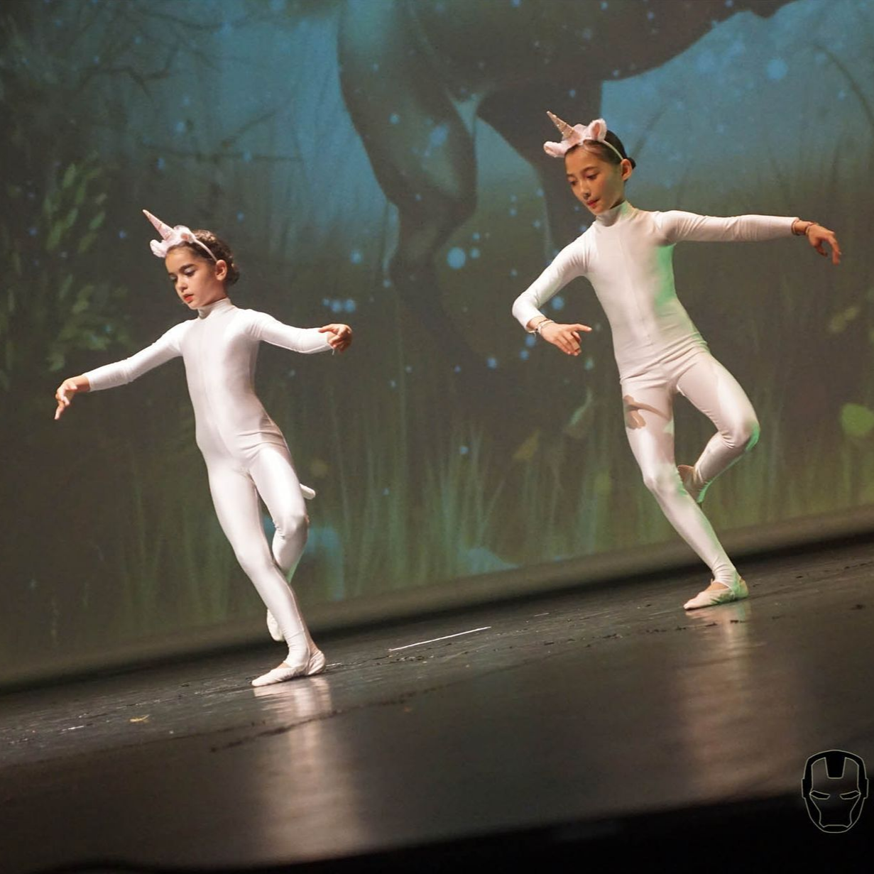 Deux enfants en costumes de licorne blanche dansent sur scène, les bras tendus. Fond vert.