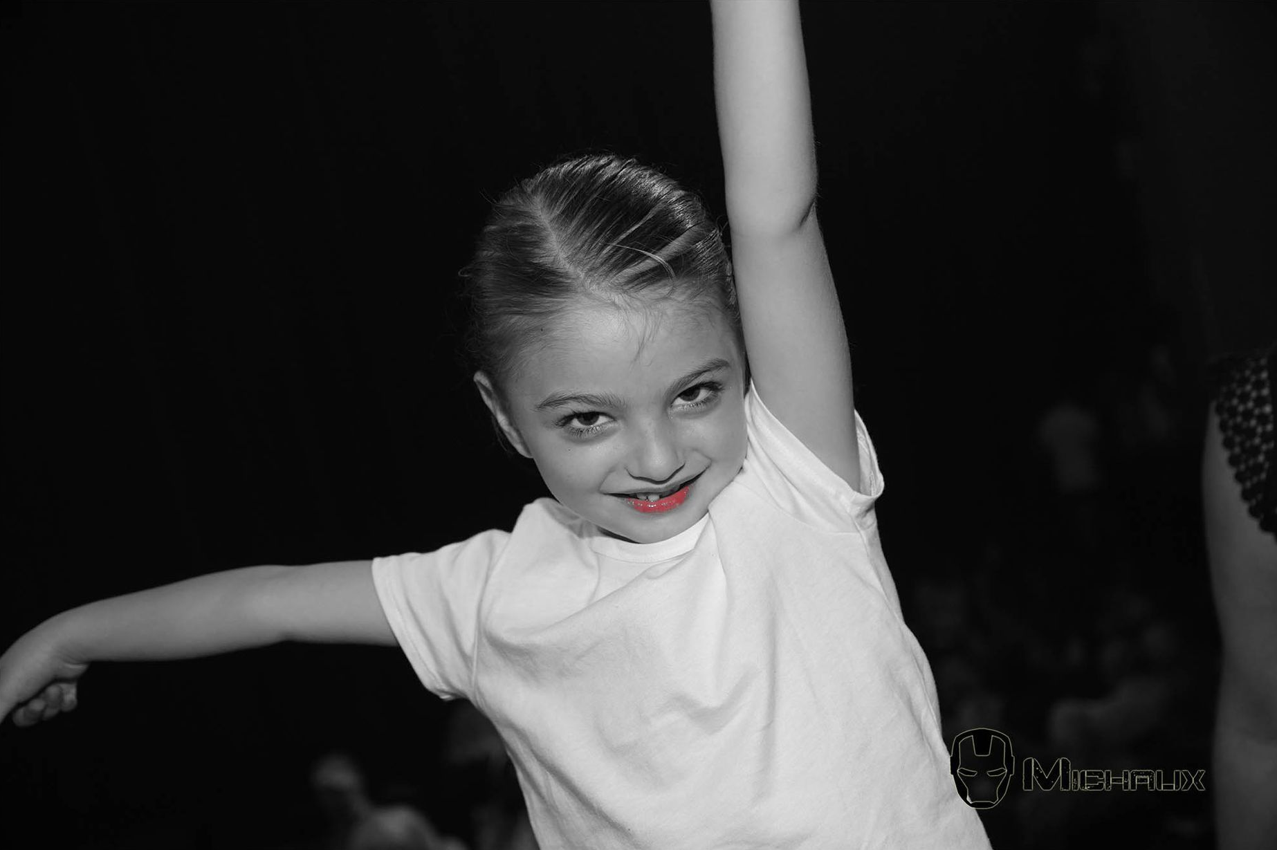 Fille en chemise blanche avec du rouge à lèvres rouge, levant le bras, souriant, dans un décor noir et blanc.