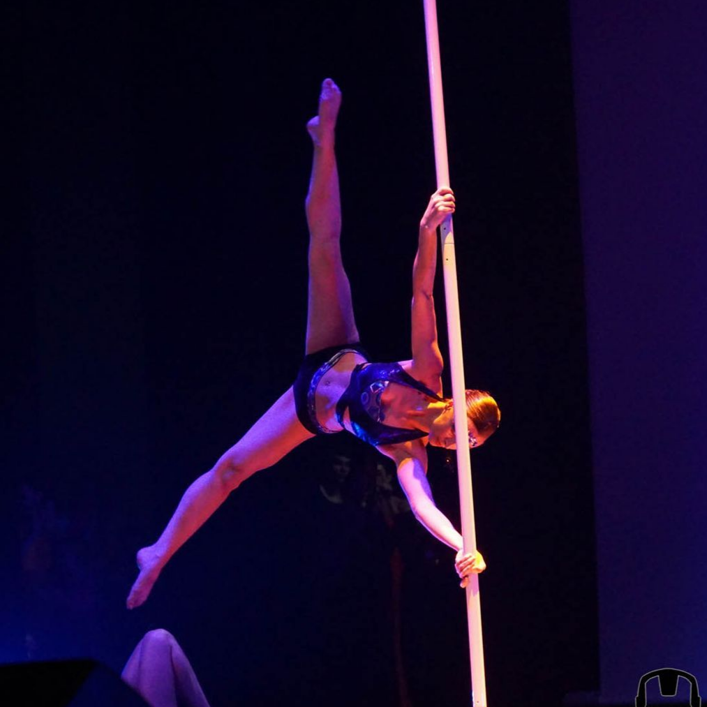 Une femme effectue un grand écart sur une barre verticale, éclairée par les projecteurs de la scène.