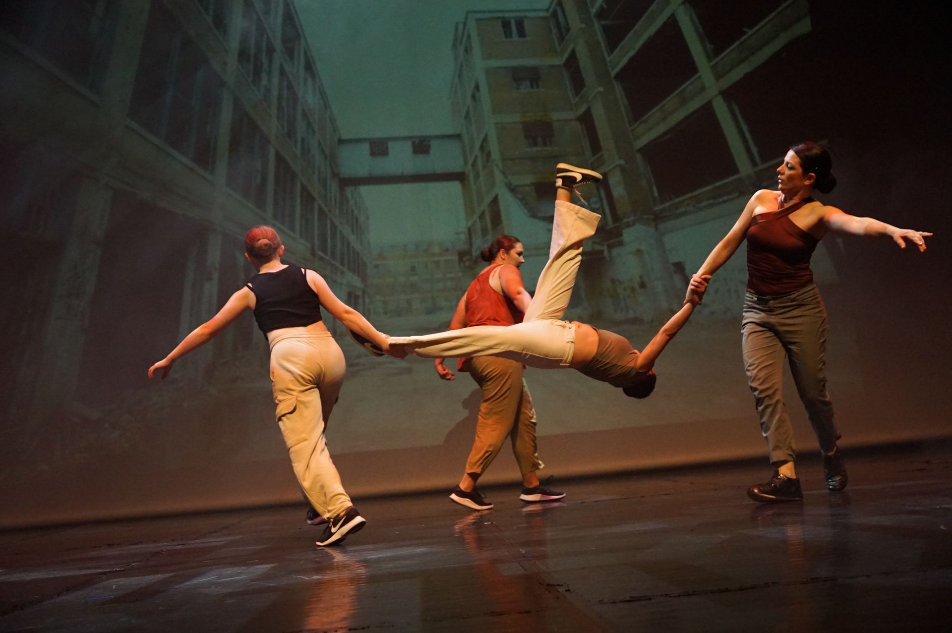 Trois danseurs évoluent sur scène ; deux d'entre eux en tiennent un troisième à bout de bras et de jambes ; le décor représente une scène urbaine.