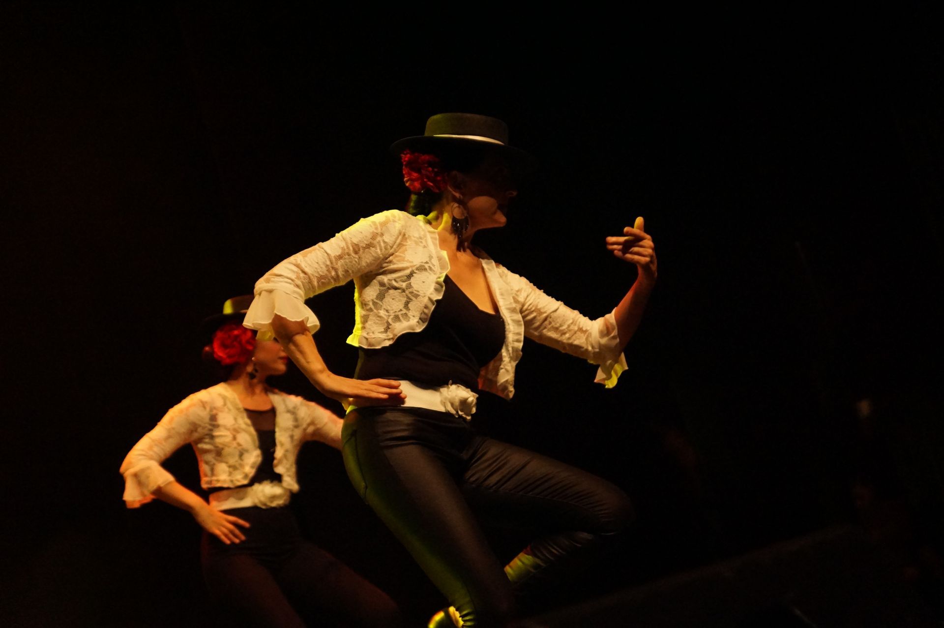 Deux danseuses de flamenco sur scène, l'une au premier plan, chapeau noir, veste blanche, pantalon noir, geste expressif du bras.