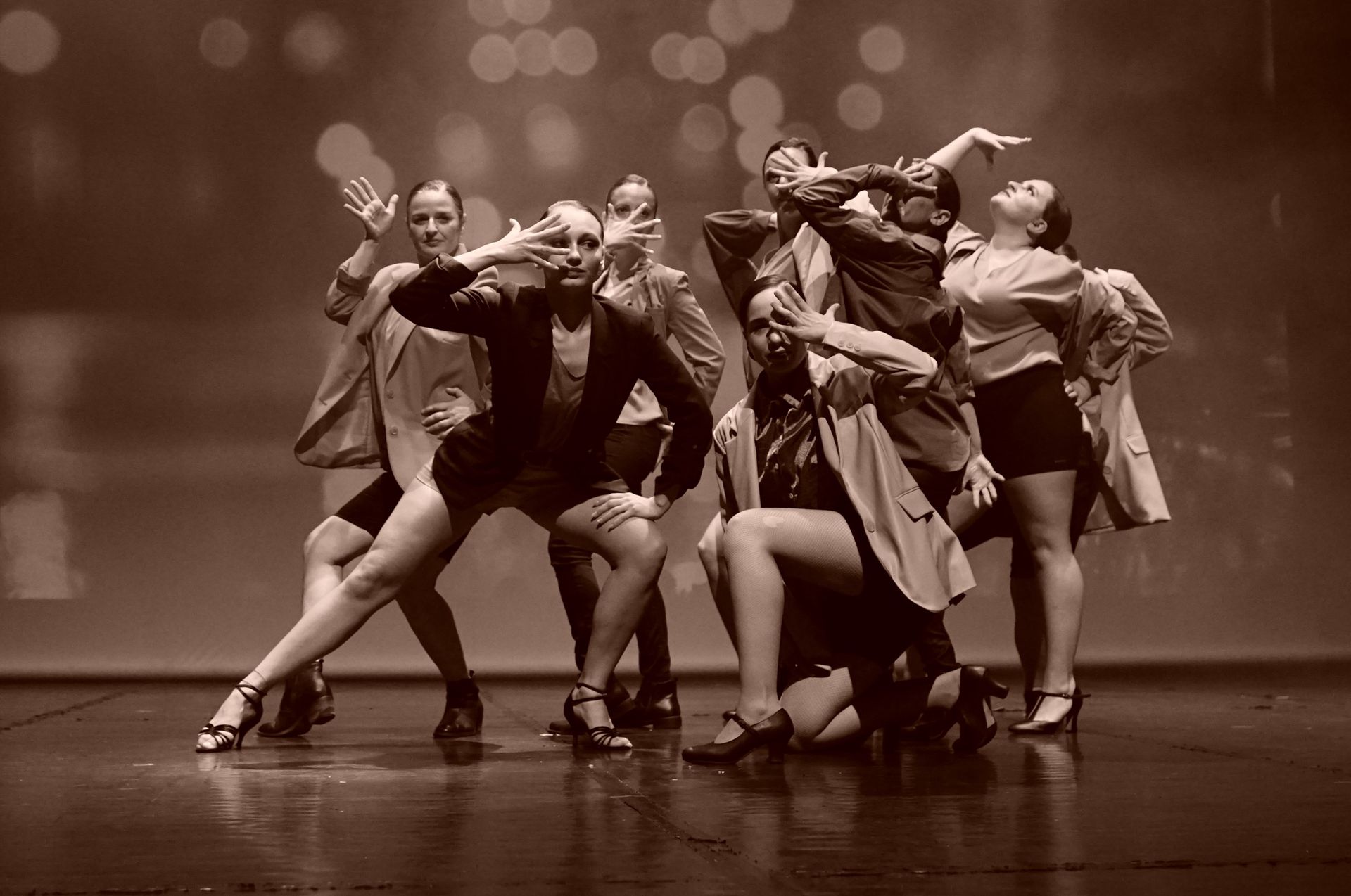 Des danseuses en groupe, se produisant sur scène. Elles portent des vestes, des shorts et des talons. Tons sépia.