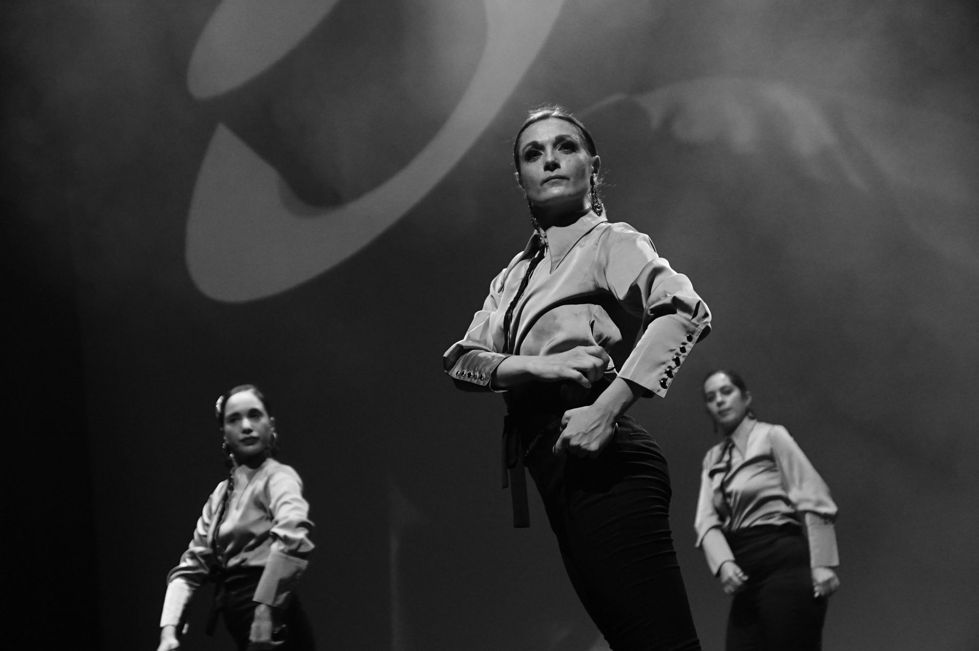 Trois danseuses en tenue de soirée se produisent sur scène. Un projecteur et un grand chapeau ornent l'arrière-plan.