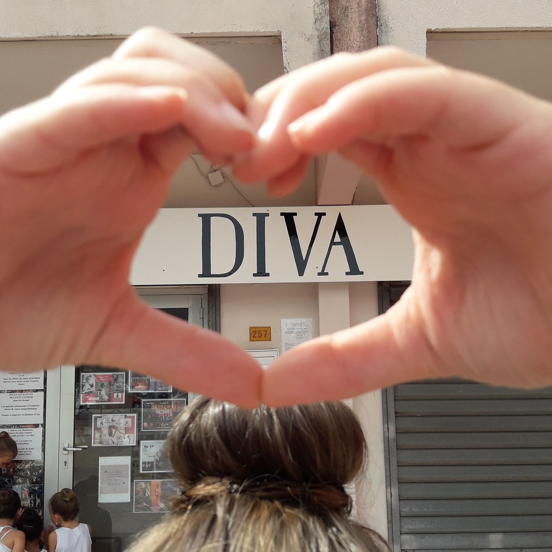 Une personne formant un cœur avec ses mains devant une enseigne de magasin sur laquelle est écrit « DIVA ».