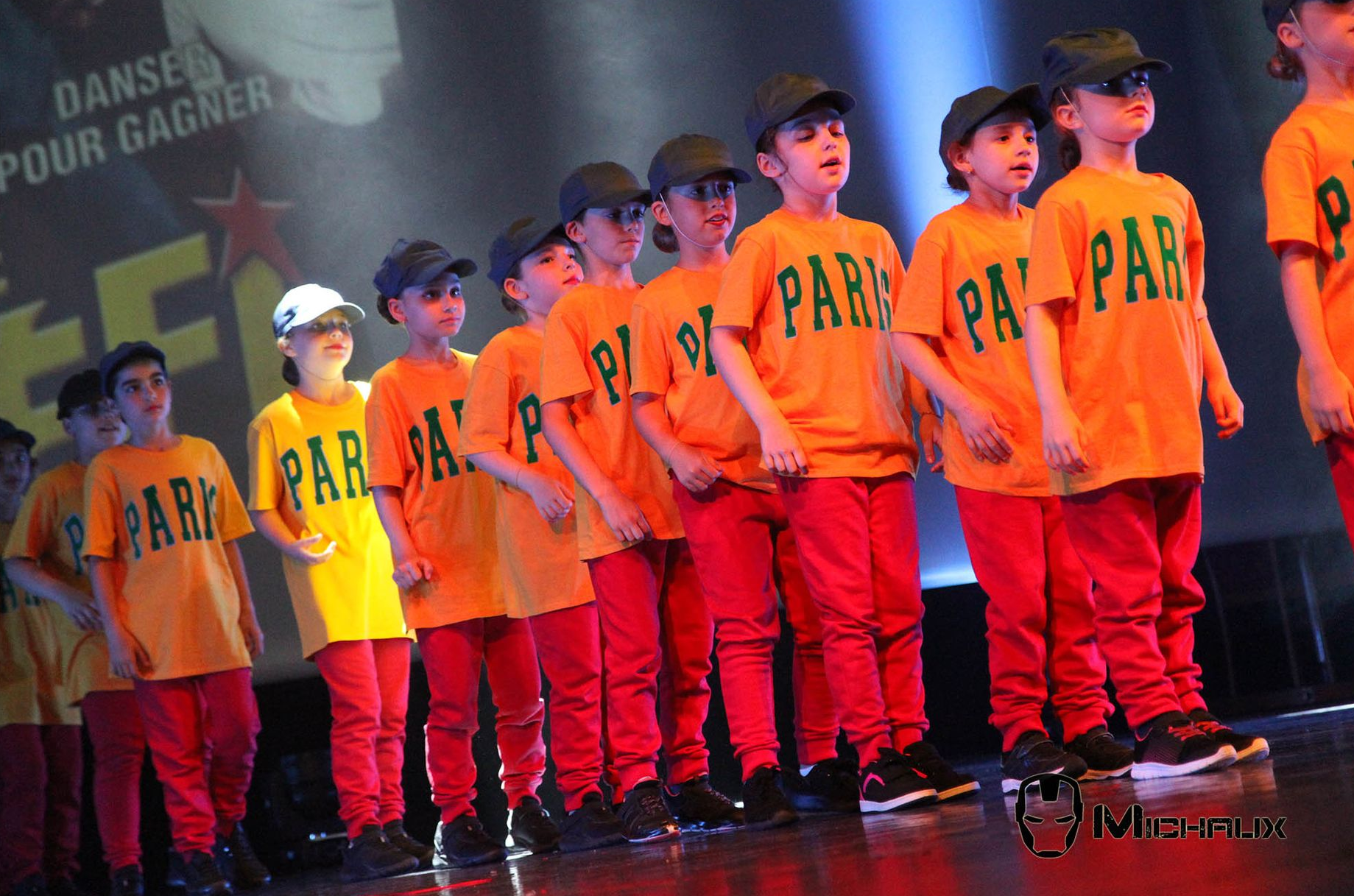 Groupe de danse d'enfants sur scène portant des chemises orange et des pantalons rouges.