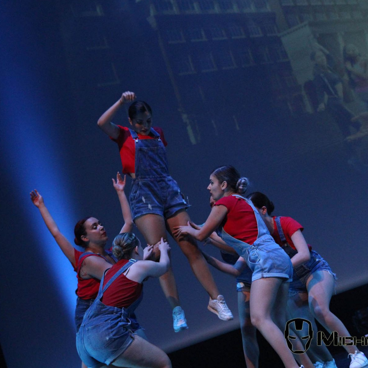 Des danseurs en hauts rouges et salopettes en jean soutiennent un danseur sautant sur scène. Fond bleu, éclairage de scène.