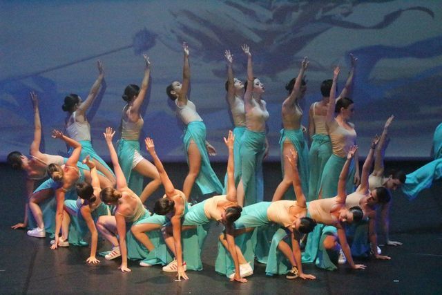 Des danseurs en costumes bleu sarcelle exécutent une chorégraphie sur scène sur fond bleu.