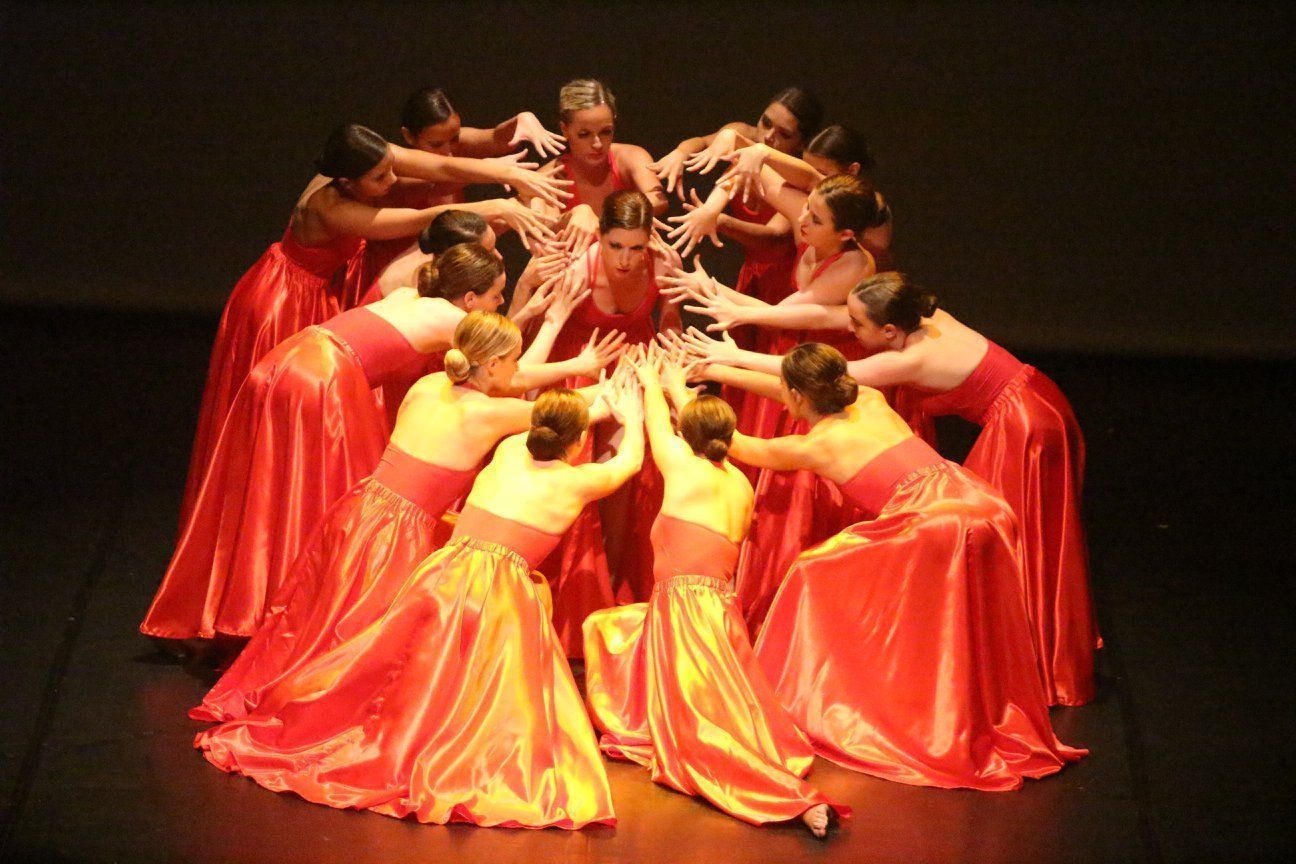 Des danseurs en longues robes rouges forment un cercle, les bras tendus vers un personnage central. Décor scénique sombre.