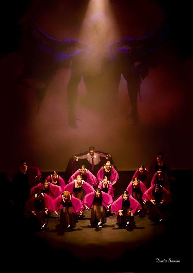 Performance sur scène : des danseurs en robes roses et costumes sombres s'inclinent devant une figure sous un projecteur.